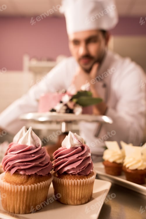 Preview: selective focus of cupcakes and confectioner standing near cake on counter in restaurant kitchen