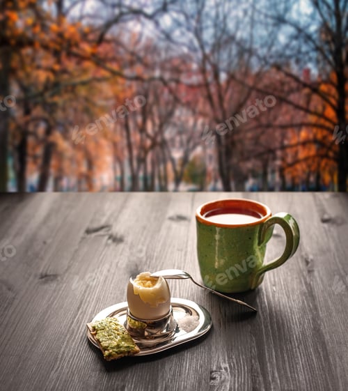 Preview: Breakfast on wooden table near by window. Boiled egg in a stand, bread with avocado and a cup of tea