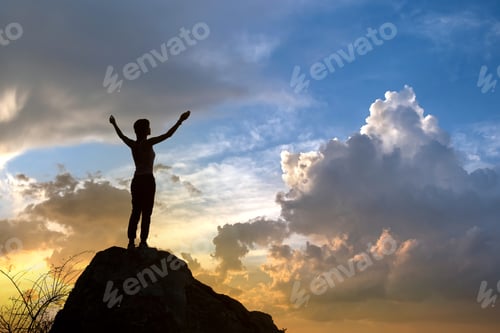 Preview: Silhouette of a woman hiker standing alone on big stone at sunset in mountains. Female tourist