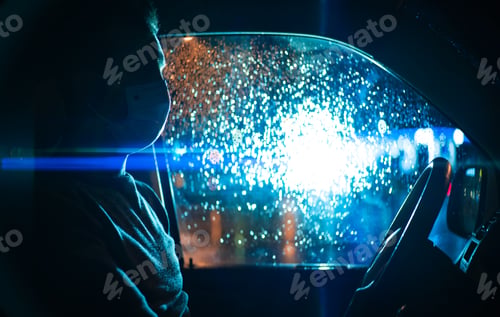Preview: The man in medical mask sits in the car on the night rainy street.