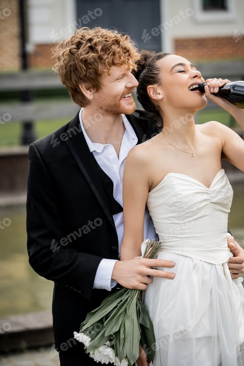 Preview: smiling redhead groom hugging african american bride drinking champagne from bottle, wedding in city