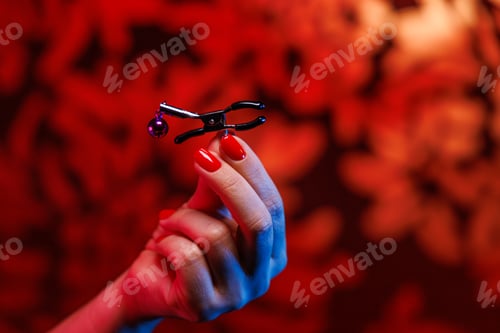 Preview: In a close up shot a woman with vibrant red nails holds a nipple clamp highlighting