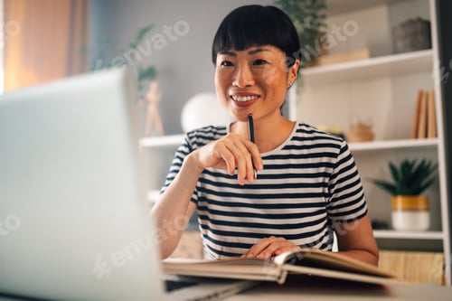 Preview: Joyful asian woman working on laptop in home office with open book