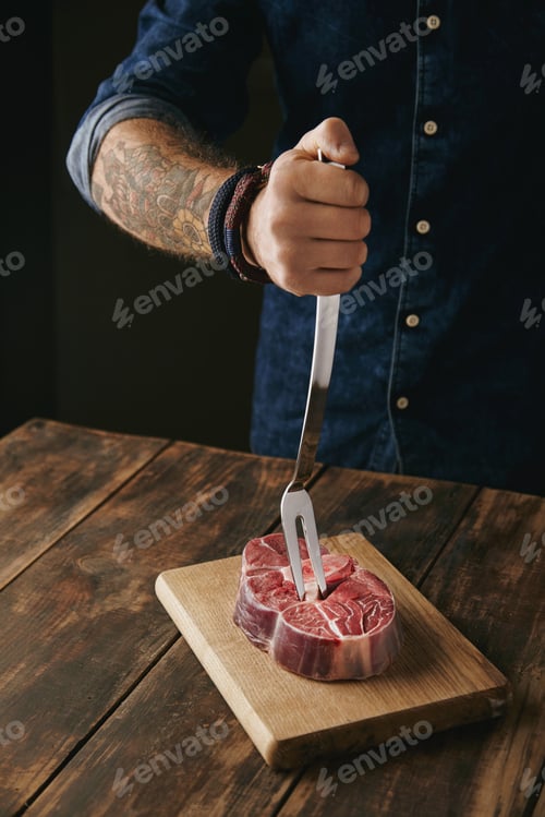Preview: butcher hand holds meat fork in raw steak