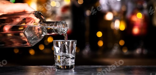 Preview: Bartender pours vodka in glass on black bar counter background with lights