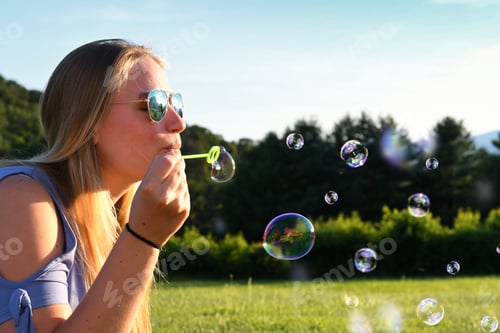 Preview: Blonde woman with aviator sunglasses blowing bubbles at golden hour on a warm summer evening.