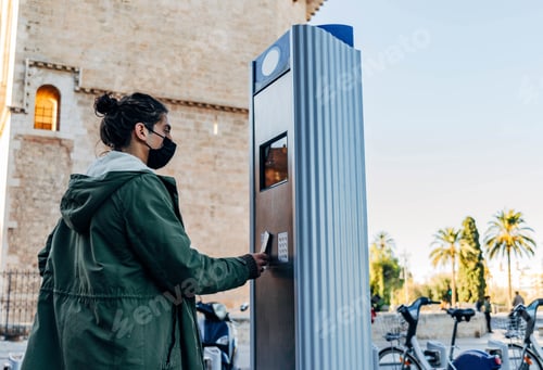 Preview: Young guy with face mask standing near a parking meter - new normal concept