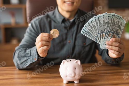 Preview: Cheerful mature japanese male show a lot of money in cash and puts bitcoin in piggy bank in office