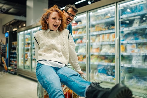 Preview: Young couple having fun while shopping in supermarket. Woman sitting in shopping cart