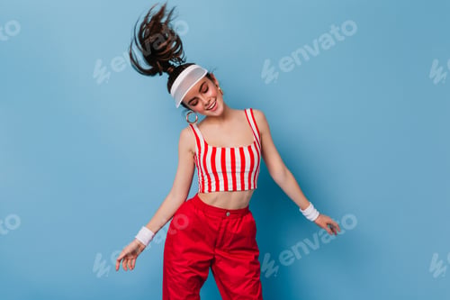 Preview: Charming woman in clothes in style of 80s dancing on blue background. Girl in cap plays hair