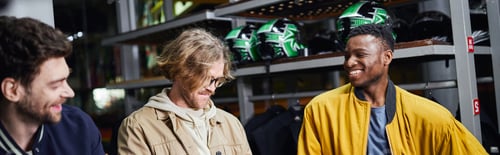 Preview: joyful interracial male friends in jackets holding helmets, indoor racing track, karting banner