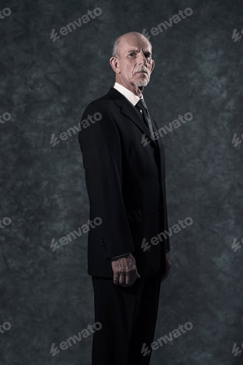 Preview: Serious Senior Businessman With Gray Beard Wearing Dark Suit And Tie. Against Grey Wall.