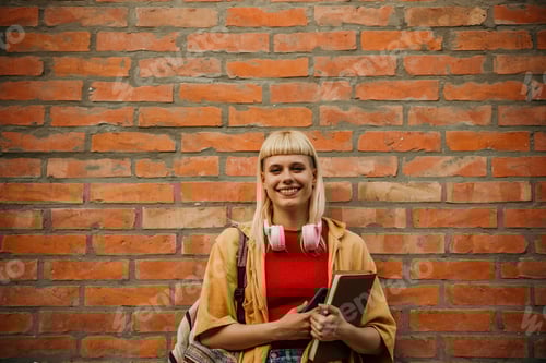 Preview: Smiling Woman with Headphones and Books near Brick Wall