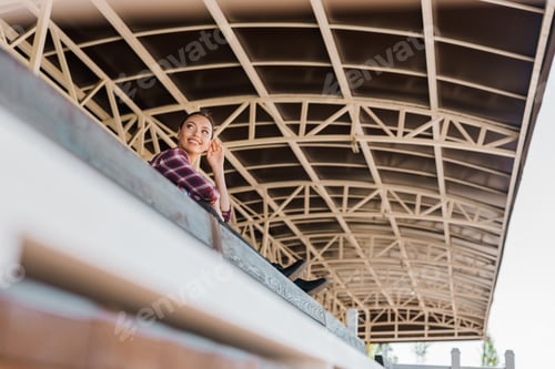 Preview: low angle view of attractive cowgirl in checkered shirt sitting on bench at ranch stadium