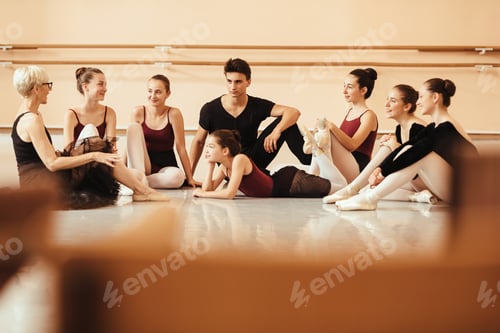 Preview: Large group of ballet dancers and their teacher talk while relaxing after rehearsal at dance studio.