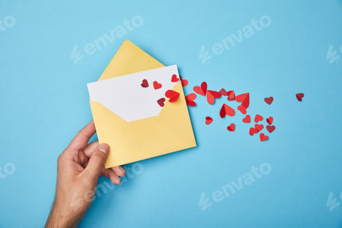 Preview: cropped view of man holding yellow envelope with blank white card and red paper hearts on blue