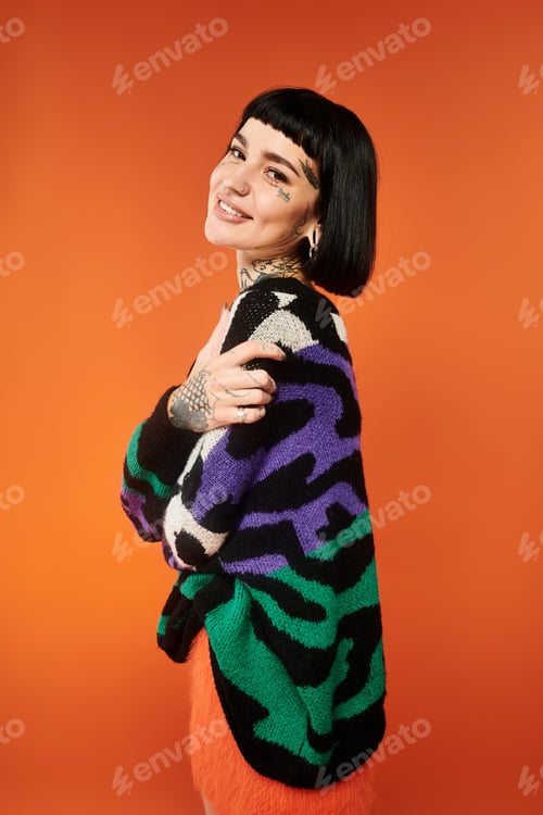 Preview: A tattooed woman poses confidently in a colorful sweater against an orange backdrop