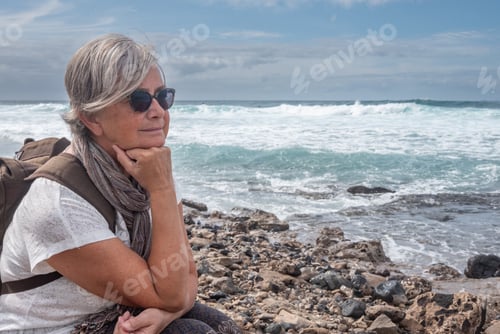 Preview: Side view of a senior woman sitting on a rocky beach and thinking and looking away.
