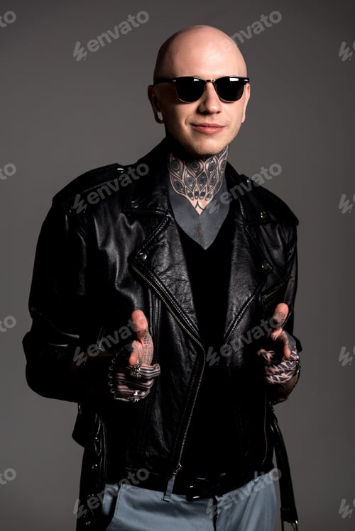 Preview: smiling tattooed man in leather jacket and sunglasses pointing at camera isolated on grey