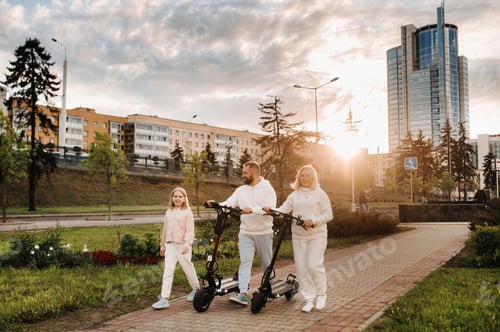 Vorschau: Eine Familie in weißen Kleidern geht bei Sonnenuntergang mit Elektrorollern durch die Stadt. Outdoor-Aktivitäten