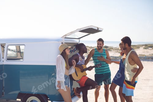 Preview: Side view of happy group of diverse friends having fun together near camper van at beach