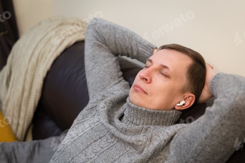 Preview: Young man relaxing on cozy couch, listening to favorite music in modern headphones.