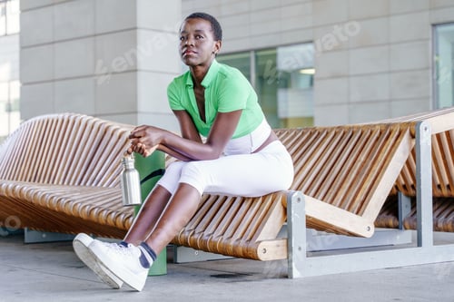 Preview: African woman with short hair sitting on wooden bench holding reusable water bottle after workout