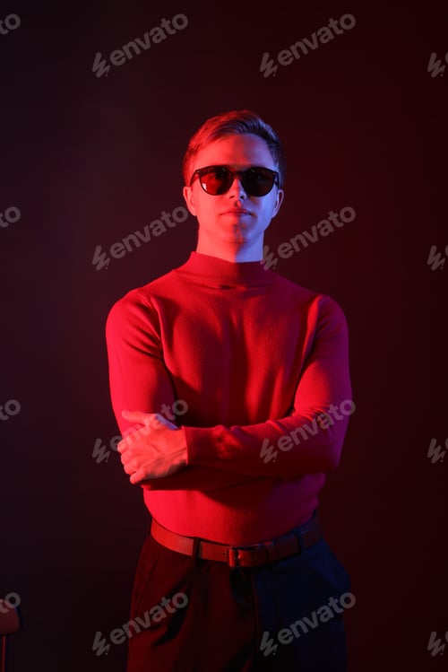 Preview: Portrait of handsome man wearing sunglasses with crossed arms in neon lights against dark background