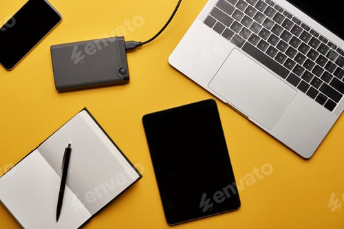 Preview: top view of various gadgets with opened notebook lying on yellow surface