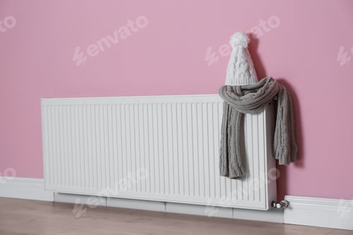 Preview: Modern radiator with knitted hat and scarf near color wall indoors. Central heating system