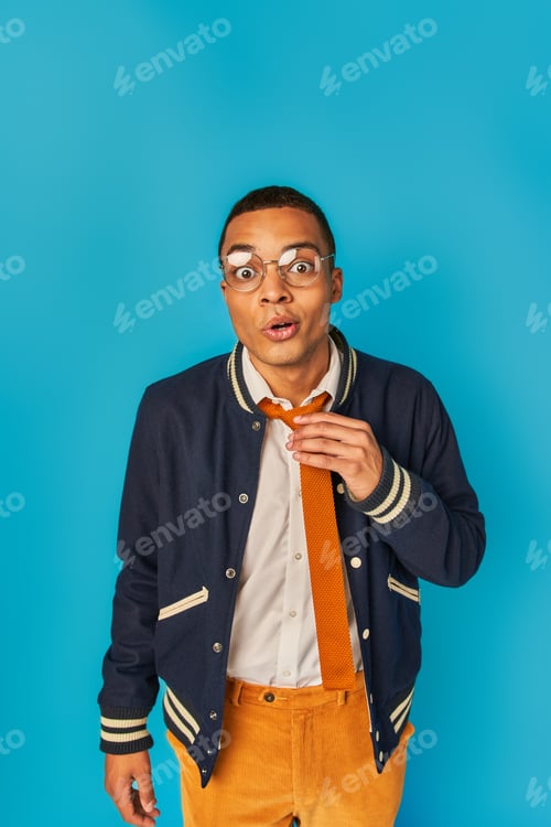 Preview: surprised and trendy african american student in jacket touching tie and looking at camera on blue