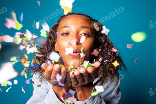Preview: Beautiful young woman studio shot blowing confetti - Isolated black female celebrating with confetti