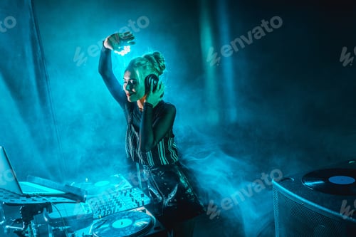 Preview: happy dj woman with blonde hair listening music in headphones in nightclub with smoke