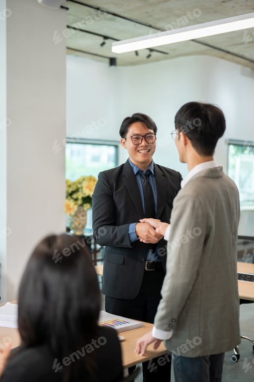 Preview: Smiling glasses businessman standing and shaking hand aside woman coworker at wooden table in office