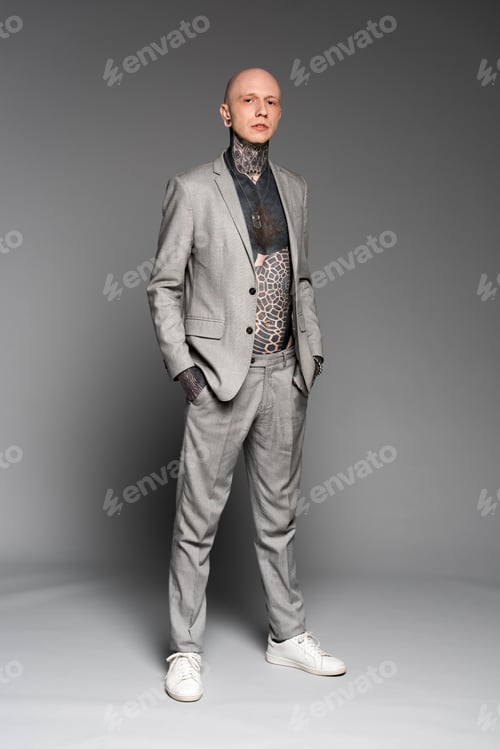 Preview: full length view of handsome stylish man with tattoos on chest standing with hands in pockets and