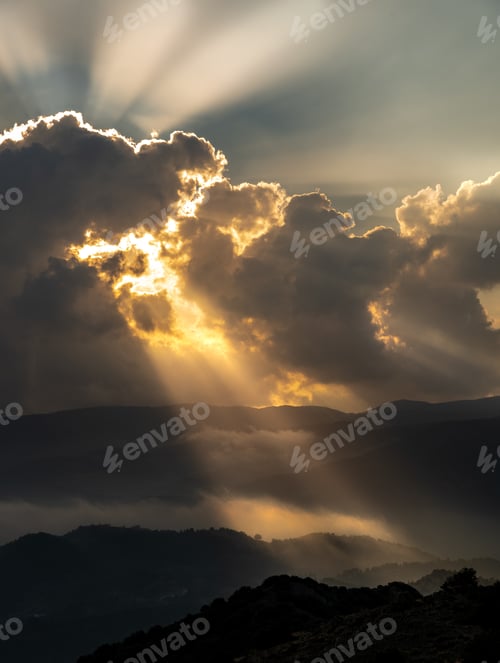 Preview: Bright sun rays light shining through dark clouds over mountain at sunset. Dramatic sky in winter