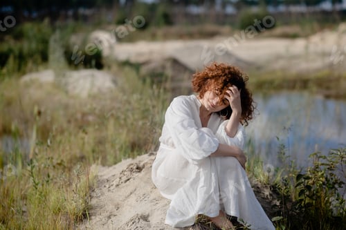 Preview: Young woman with curly hair and wearing white dress is standing by the lake