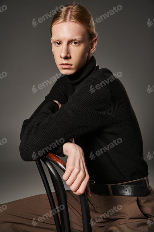 Preview: good looking non binary model in stylish attire with red hair sitting on chair and looking at camera
