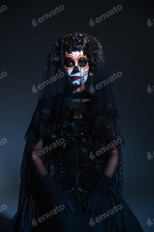 Preview: front view of woman in black costume and traditional santa muerte makeup sitting on dark blue