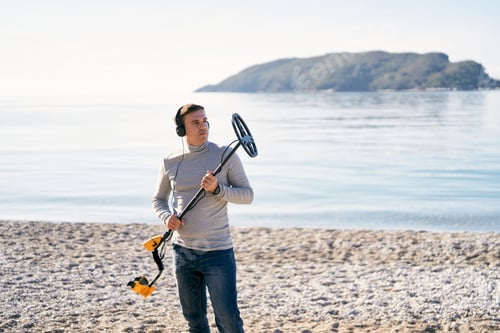 Preview: Young man stands on the seashore holding up a metal detector