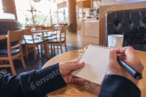 Preview: Closeup image of a hand holding and writing on blank notebook on table in cafe