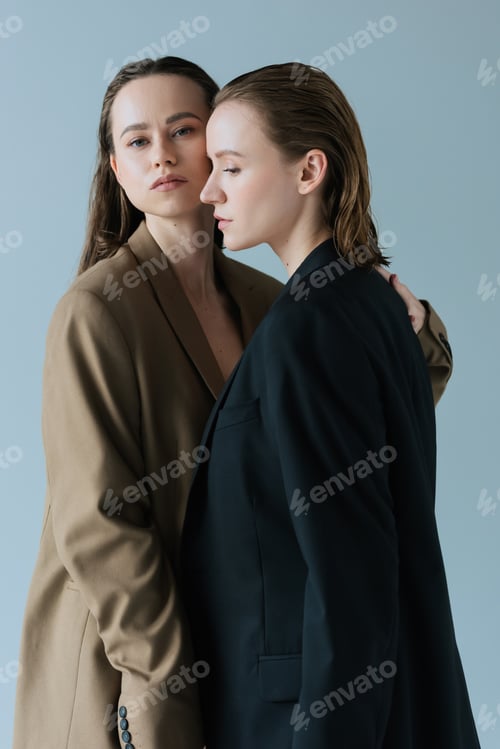 Preview: young woman in beige blazer hugging lesbian partner and looking at camera isolated on grey