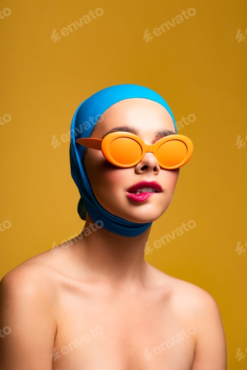 Preview: fashionable naked girl in scarf and orange sunglasses biting lip, on yellow