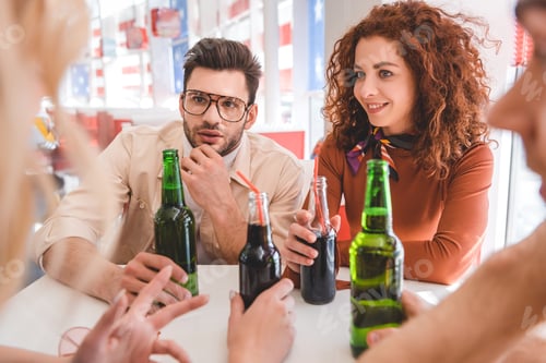 Preview: selective focus of handsome and attractive friends holding glass bottles with drink and talking at