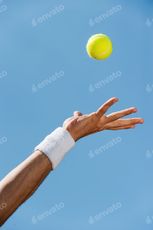 Preview: Serving ball. Close-up of male hand in wristband throwing tennis ball against blue sky