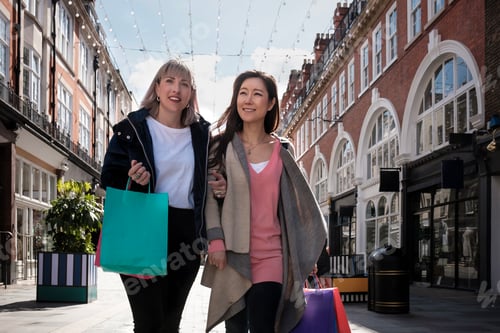 Preview: Portrait of two multiracial mature women walking along a London street wearing shopping bags.