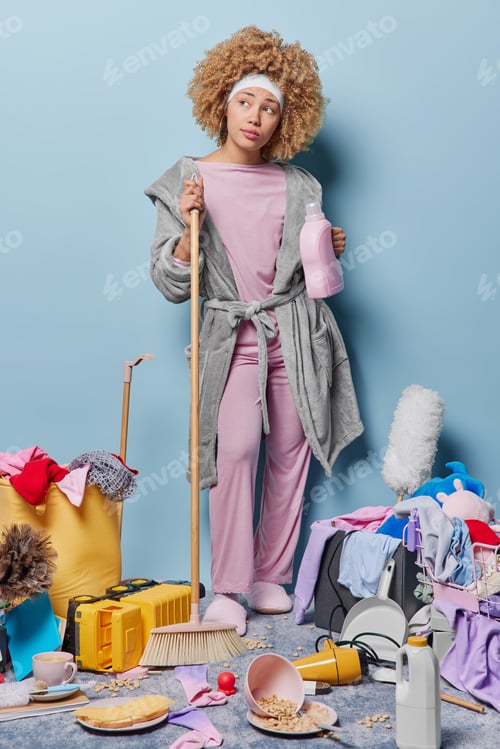 Preview: Thoughtful woman with curly hair wears domestic robe anss headband poses with brush and bottle of