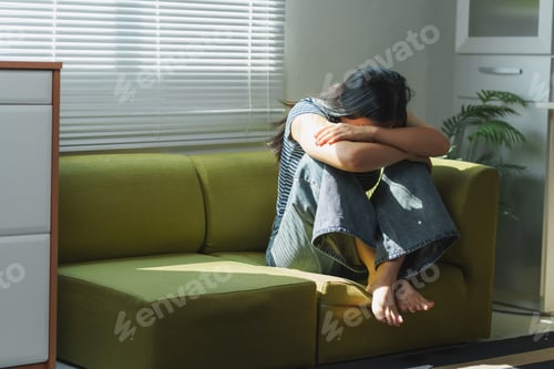 Preview: Sad young woman sitting on sofa holding head in hands