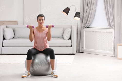 Preview: Young woman doing exercise with dumbbells on fitness ball at home. Space for text