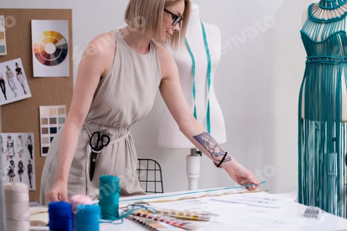 Preview: Smiling tailor measures green thread with tape on table in fashion studio woman designer work